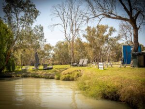 Grassy riverbank campsite with deck chairs and scattered trees near calm water.