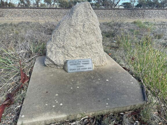 Stone memorial with plaque set on a concrete base in a grassy roadside area.