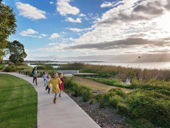 Children running along a lakeside path under a cloudy blue sky.