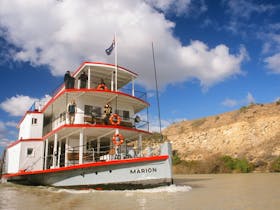 Paddle steamer with passengers cruising on a calm river under a clear blue sky.