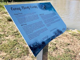 Informational sign titled 'Ferries, Floods & Loo's' beside a calm riverside.