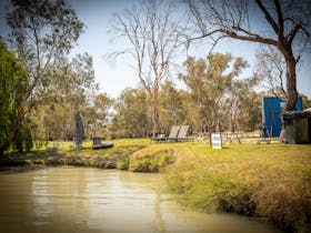 Grassy riverbank campsite with deck chairs and scattered trees near calm water.
