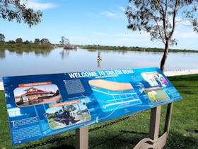 Welcome sign at Tailem Bend overlooking a calm river with trees and blue sky.